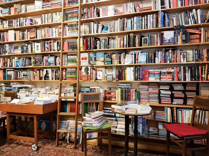 Shakespeare and Company Booksellers interior, a large bookshelf with ladder, and a few tables, stools, and chairs covered with books.