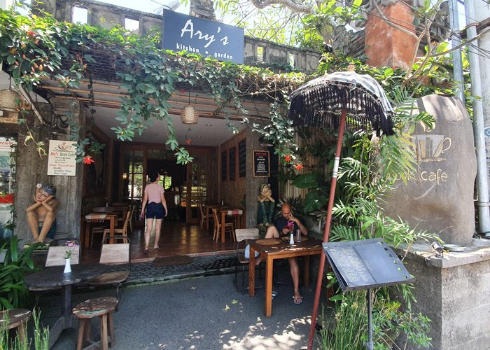 Exterior of Ary's Book Cafe in Ubud, Bali, Indonesia.  A stone builing with a large open entrance covered by green vines. Wooden tables and chairs line both sides of the space.