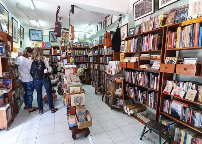 Interior of Ganesha Bookshop in Ubud, Bali, Indonesia. Many wooden shelves are filled with books, 3 guitars hang from the ceiling and three people are browsing on the left. 