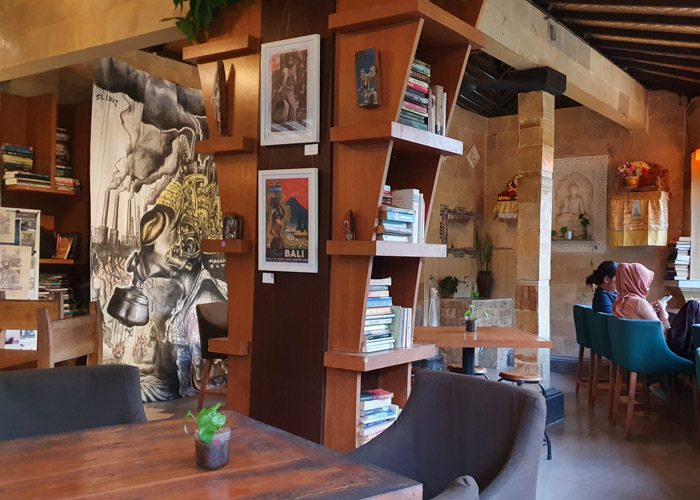 Interior of Little Talks book cafe in Ubud, Bali, Indonesia. Books are stacked on wooden shelves, a large black and white artwork hangs on the rear wall. Two women are sitting in green chairs on the right, under a small alter.