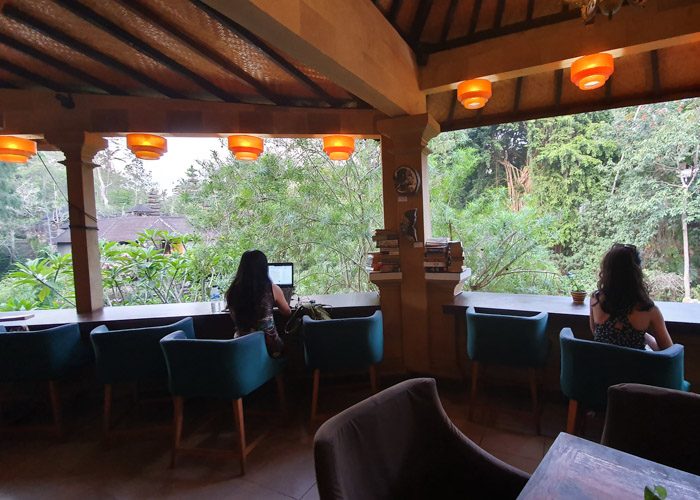 Interior of Little Talks book cafe in Ubud, Bali, Indonesia. Two women sit in green chairs, facing a beautiful view of green trees and a temple in the distance.