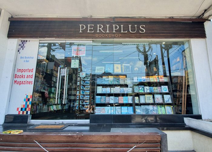 Exterior of Periplus Bookshop in Ubud, Bali, Indonesia. A simple glass fronted shop, with neatly arranged books visible through the window. A wooden sign above the glass says Periplus Bookshop, and a white sign on the left says 'Imported Books and Magazines'.