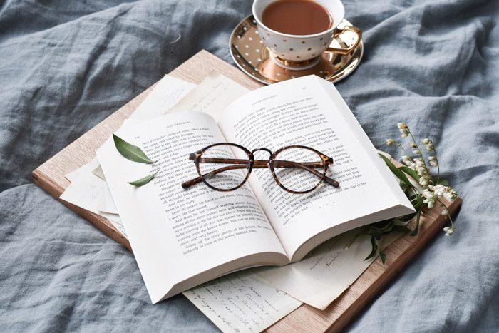 How to set reading goals for the new year: photo representing reflection. A pair of glasses sit on an open book on blue linen sheets, next to tea in a polka dot teacup.