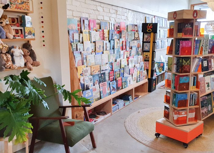 Interior of Squishy Minnie Bookstore in Kyneton. Childrens picture books are displayed on a large shelf with their covers facing forwards. A shelf on the left displays stuffed animal toys above a green indoor plant and a green armchair.