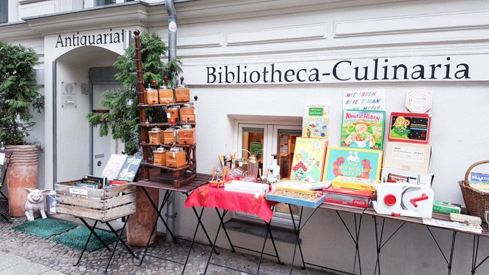 White facade of this bookshop with a few tables selling a mixture of books, kitchen equipment, and other assorted small items.