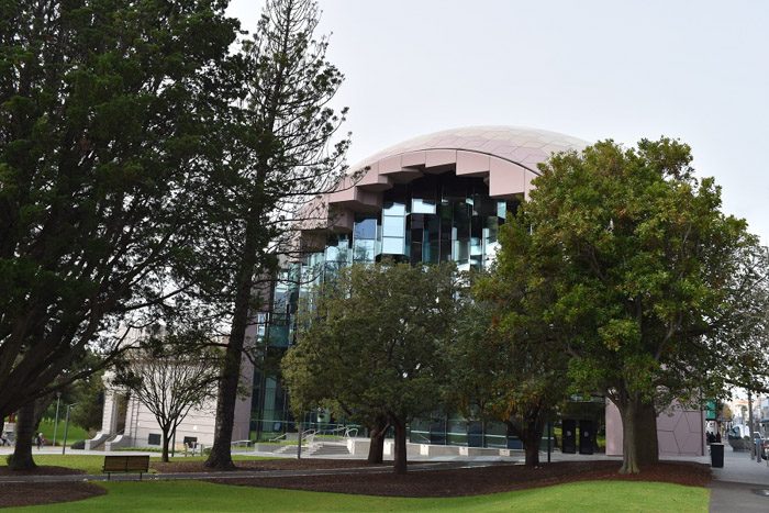 Geelong Library exterior, a domed pale pink building with geometric windows is just visible behind large green trees in a park.