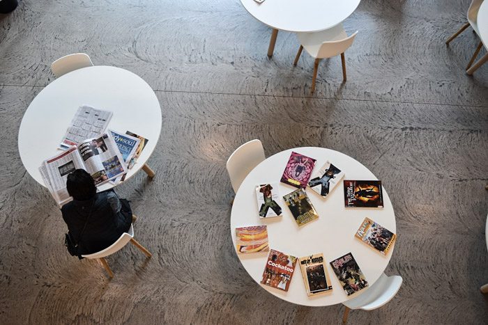 Geelong Library interior, round white tables are seen from above. A person sits at one table reading a newspaper, another table has an array of graphic novels spread on it.