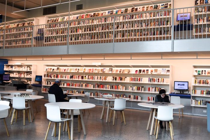 Geelong Library interior, long white shelves are stacked neatly with books on two levels, the upper level with a balcony. Two people sit at white tables in the foreground.