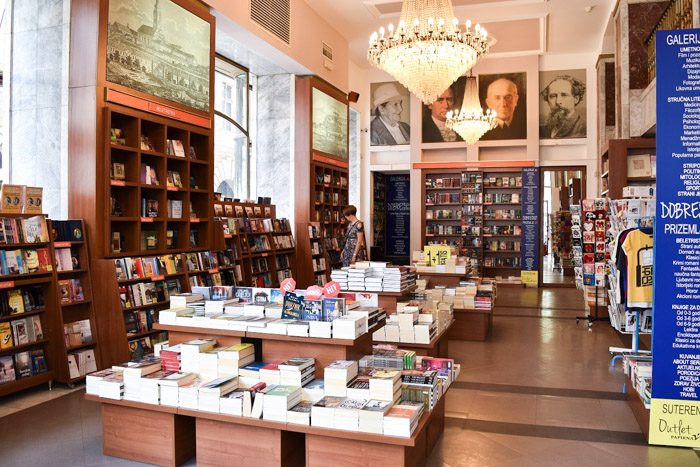 Interior of Knjizara Akademija. Two large chandeliers light a large room filled with bookshelves and tables of books. Book Lover's Guide to Belgrade, Serbia.